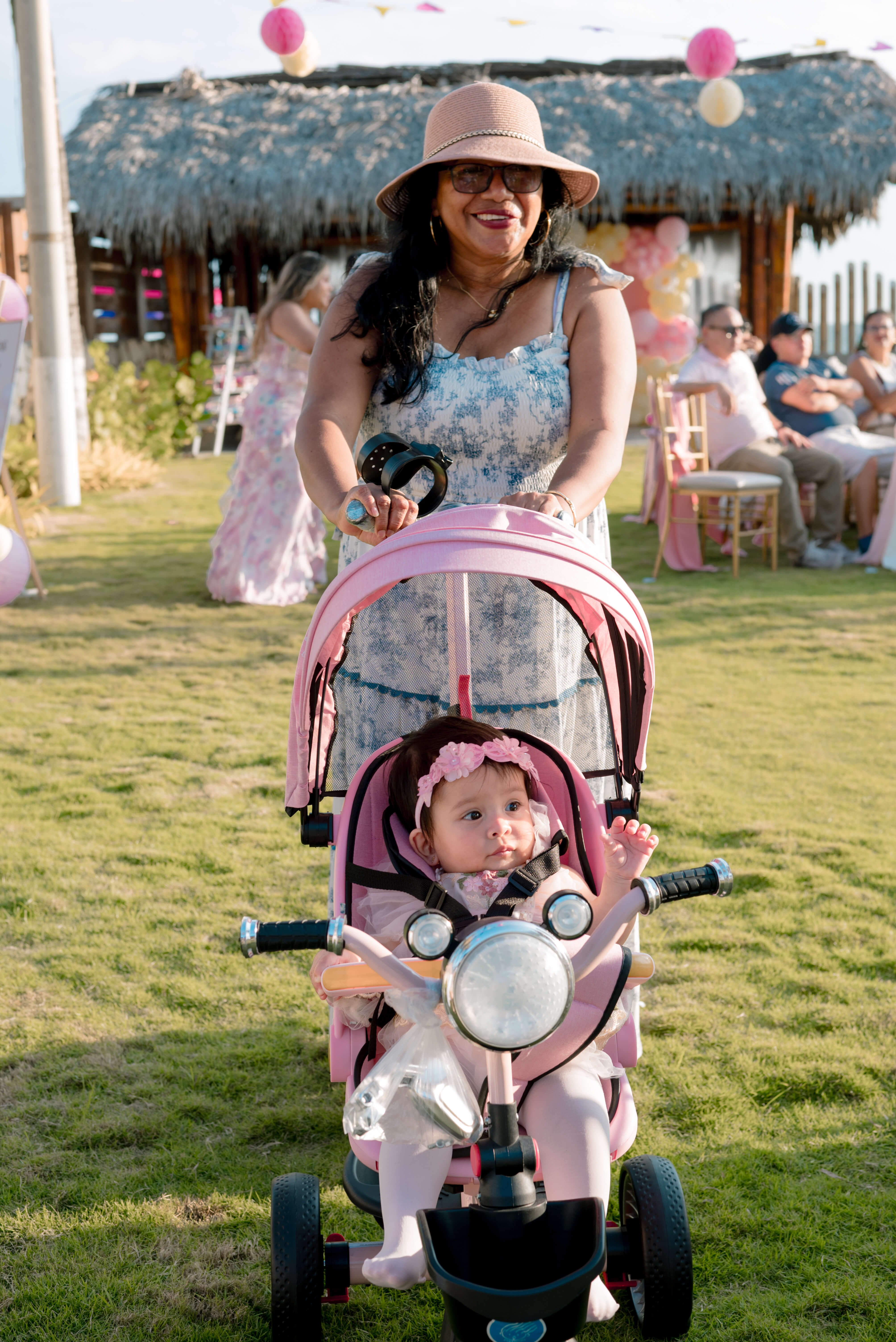 Bebé Mía sentada en su tricibici rosada empujada por su abuelita durante la celebración de su primer cumpleaños en Las Marías Playas